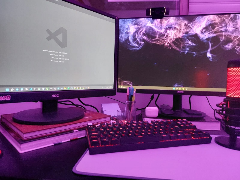 Imagem de uma mesa preta com teclado com luzes vermelhas, dois monitores ligados com tela de fundo e VS Code aberto e objetos de papelaria ao redor.